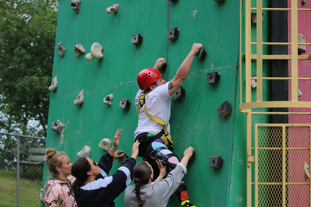 Person with red helmet climbing a climbing wall with 3 people below.